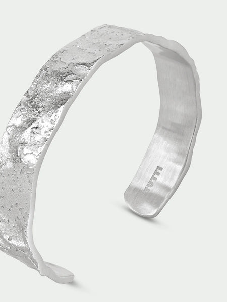 Tutti & Co Solstice Bangle in Silver A wide silver cuff bracelet with a heavily textured, hammered metallic exterior and a brushed silver interior, shown on a light gray background.
