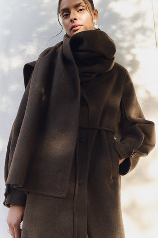 Levete Room Owa 33 Jacket in Coffee Melang Woman wearing a brown coat with a scarf against a blurred natural background