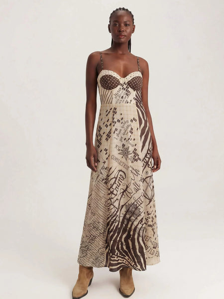 Me369 Muse Corset Dress in Noir Muse A model wearing a sleeveless maxi dress with a beige and brown abstract print, featuring a sheer polka-dot bustier bodice and thin straps, paired with tan ankle boots, against a plain light background.
