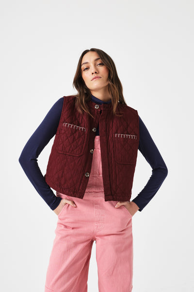 Seventy + Mochi Quilted Pablo Vest in Mulberry Woman wearing a maroon vest over a navy long-sleeve shirt with pink pants on a white background