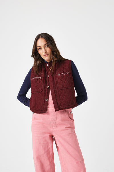 Seventy + Mochi Quilted Pablo Vest in Mulberry Woman wearing a maroon vest over a blue long-sleeve shirt and pink pants on a white background