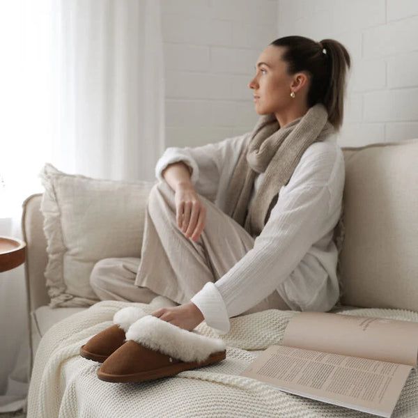 Emu Australia Jolie in Chestnut W10015 Woman sitting on a couch wearing a white sweater, beige pants, and brown slippers with fur trim.