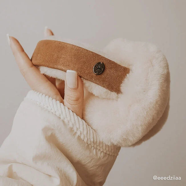 Emu Australia Angahook Earmuffs in Chestnut W9403 Close-up of a hand holding a fluffy white slipper with a brown strap and button on a neutral background.