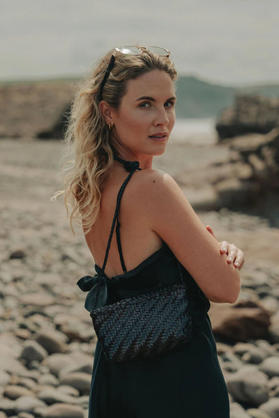 Ellyla Sia Crossbody Woven Bag in Black Woman in a black dress standing on a rocky beach with mountains in the background
