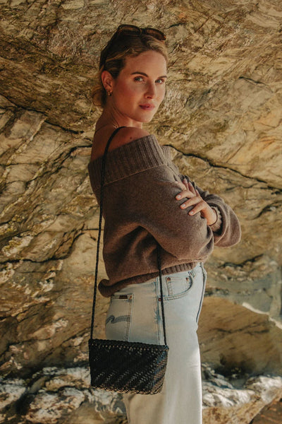 Ellyla Sia Crossbody Woven Bag in Black Woman wearing a brown off-shoulder sweater and light blue jeans against a stone wall.
