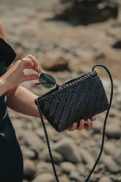 Ellyla Sia Crossbody Woven Bag in Black Person holding a woven handbag with sunglasses on a rocky surface