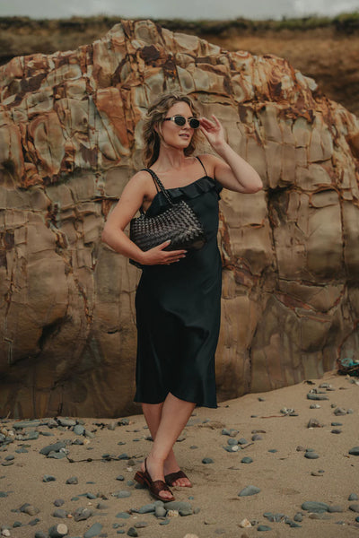 Ellyla Selina Two Long Handles Woven Bag in Espresso Woman in a black dress standing on a rocky beach with large rocks in the background