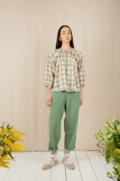 Bonte Florette Top in Clover Woman wearing a patterned blouse and green pants standing in front of a beige wall with floral decorations.