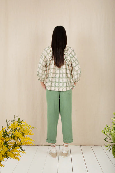 Bonte Florette Top in Clover Person wearing a patterned shirt and green pants standing against a beige wall with yellow flowers on either side.