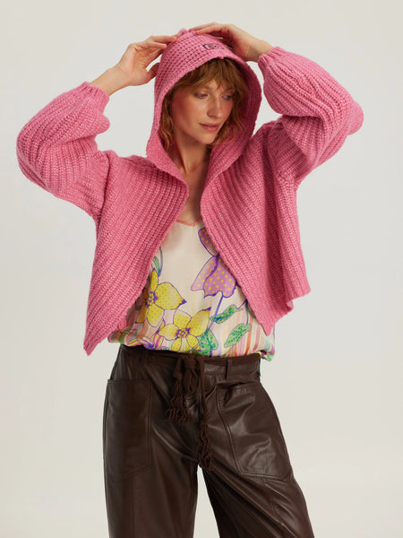 Me369 Romi Hoodie in Fuchsia A woman wearing a chunky pink knit cropped hoodie jacket over a floral top and dark brown pants, posing against a light gray studio background.